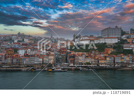 View of Porto, Portugal, featuring the colorful Ribeira district, Clerigos Tower, and Episcopal Palace. Traditional Rabelo boats dot the Douro River under dramatic clouds. 118719891