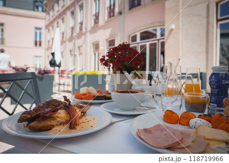 Experience a lavish breakfast spread featuring eggs benedict, cold cuts, fresh fruit, and pastries at a sophisticated hotel with European architecture and lush greenery. 118719966