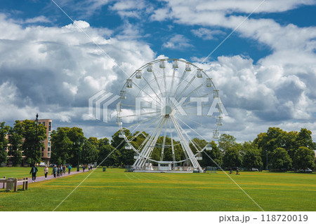 A spacious park in Cambridge, UK, featuring a prominent Ferris wheel, lush trees, and a partly cloudy sky. People walk along a pathway with benches. A spacious park in Cambridge, UK, featuring a prominent Ferris wheel, lush trees, and a partly cloudy sky. People walk along a pathway with benches. 118720019