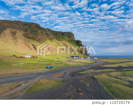 A stunning aerial view of Iceland's rugged coastline featuring a winding dirt road, green cliffside with small houses, and the vast ocean under a bright blue sky. A stunning aerial view of Iceland's rugged coastline featuring a winding dirt road, green cliffside with small houses, and the vast ocean under a bright blue sky. 118720179
