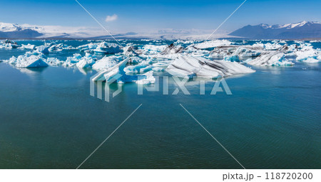 Stunning aerial view of a deep blue glacial lagoon in Iceland, featuring scattered white and blue icebergs and snow-covered mountains in the background. 118720200
