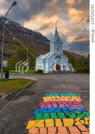 The picturesque blue Seydisfjordur Church in Iceland, set against snow-capped mountains and a dramatic sky, with a vibrant rainbow cobblestone path leading up to it. 118720242