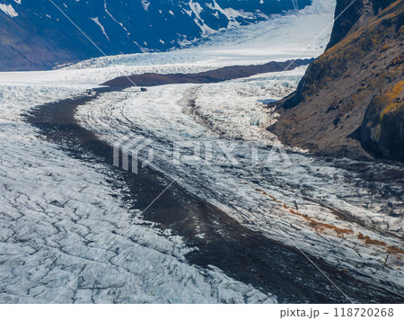 Stunning aerial view of a glacier in Iceland, showcasing snow and ice, a dark rocky ridge, and surrounding mountains under a clear blue sky. 118720268