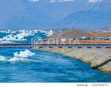A metal bridge with a wooden walkway spans a bright blue river with floating icebergs and a backdrop of snow-covered mountains in Iceland. A metal bridge with a wooden walkway spans a bright blue river with floating icebergs and a backdrop of snow-covered mountains in Iceland. 118720271