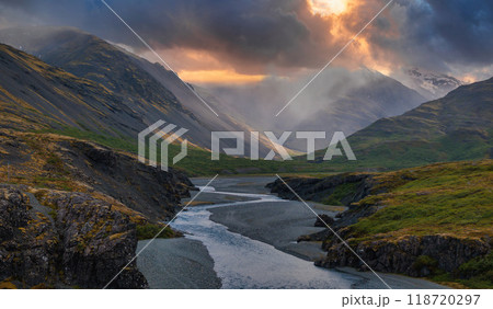 A light blue river winds through a valley in Iceland, surrounded by dark gray mountains and dramatic clouds with a bright orange glow breaking through. 118720297