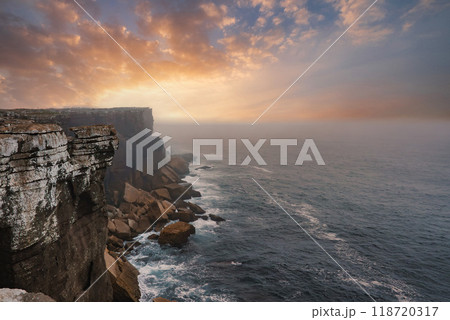 Breathtaking view of the western coast of Portugal featuring dramatic cliffs, vibrant sunset hues, and misty ocean spray. Waves crash against the rugged rocks. 118720317