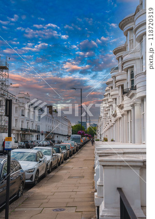 A serene urban scene in London, UK, featuring classic white terraced houses, parked cars, and a vibrant sky at dusk. Pedestrians and scaffolding add life and detail. A serene urban scene in London, UK, featuring classic white terraced houses, parked cars, and a vibrant sky at dusk. Pedestrians and scaffolding add life and detail. 118720319