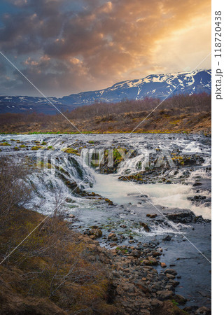 A stunning waterfall in Iceland flows over rocky terrain with snow-capped mountains in the background. The dramatic sky features dark clouds and a warm, glowing sunset. 118720438