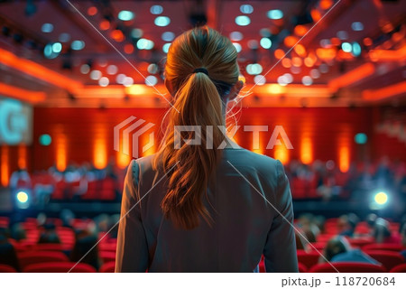 An individual looks toward the stage in a brightly lit auditorium, as attendees gather for a business conference focused on networking and learning 118720684