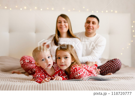 The children in Christmas pajamas are reading a book, while their parents embrace in the background. The children in Christmas pajamas are reading a book, while their parents embrace in the background. 118720777