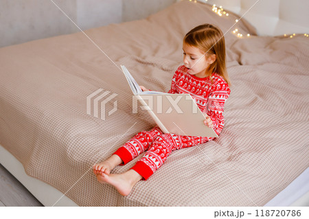A girl in a Christmas pajama is reading a large book on the bed 118720786
