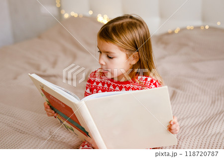 The girl in festive pajamas is lying on the bed and reading a big book 118720787