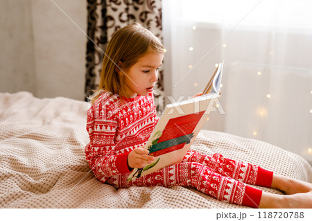 A little girl wearing holiday sleepwear is on the bed, reading a big book 118720788