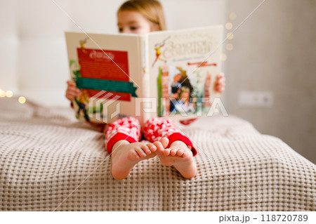 A girl in a Christmas pajama is reading a large book on the bed, focusing on her feet 118720789