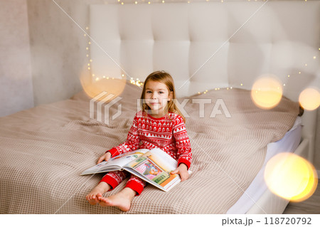 The girl in red Christmas pajamas is sitting on the bed and enjoying a big book 118720792