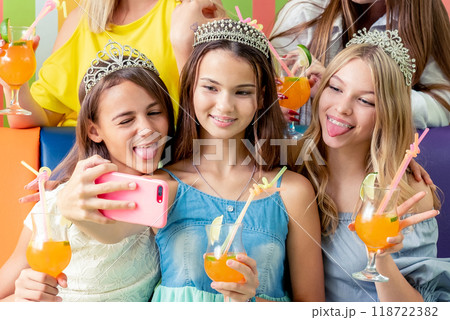 Pretty smiling teenage girls in dresses and crowns sit hugging together holding beverages and taking selfie at birthday party 118722382