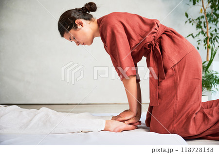 Masseuse making thai yoga massage. Woman therapist doing traditional massage treatment, stretching female patient's body. Client lying on mat, enjoying hand massage in calm atmosphere. Masseuse making thai yoga massage. Woman therapist doing traditional massage treatment, stretching female patient's body. Client lying on mat, enjoying hand massage in calm atmosphere. 118728138
