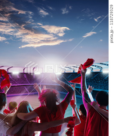 Group of fans in red shirts celebrating under colorful twilight sky, waving flags and scarves as field bathed in soft evening light. 118729329