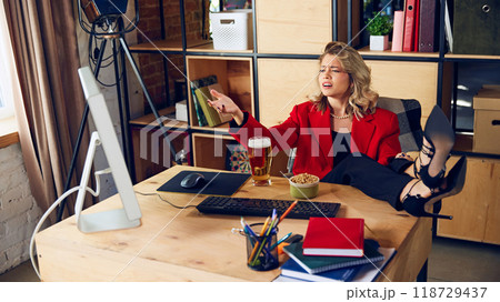 Beer in hand, feet on desk, young woman points at screen, seemingly frustrated while multitasking between work and casual drink. Beer in hand, feet on desk, young woman points at screen, seemingly frustrated while multitasking between work and casual drink. 118729437