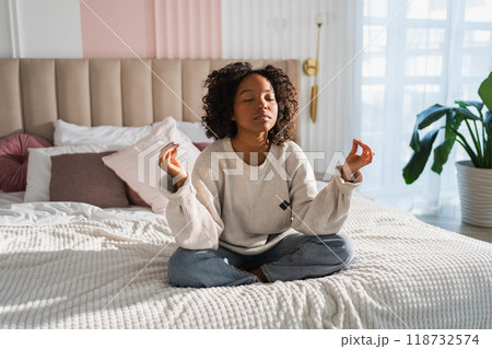 Yoga mindfulness meditation. Young healthy African girl practicing yoga at home. Woman sitting in lotus pose on bed meditating smiling relaxing indoor. Girl doing breathing practice. Yoga at home Yoga mindfulness meditation. Young healthy African girl practicing yoga at home. Woman sitting in lotus pose on bed meditating smiling relaxing indoor. Girl doing breathing practice. Yoga at home 118732574