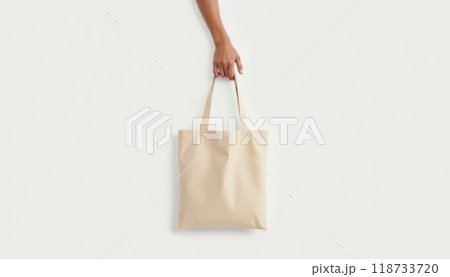 A person holds a plain beige shopping bag by the handle against a white background. The scene is minimalistic and focused on the bag. A person holds a plain beige shopping bag by the handle against a white background. The scene is minimalistic and focused on the bag. 118733720