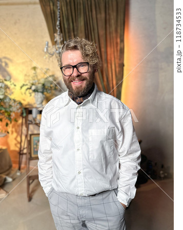 Cheerful millennial man in a white shirt smiles warmly in a cozy, classically decorated room with soft lighting. Joy, relaxation and timeless elegance concept 118734533