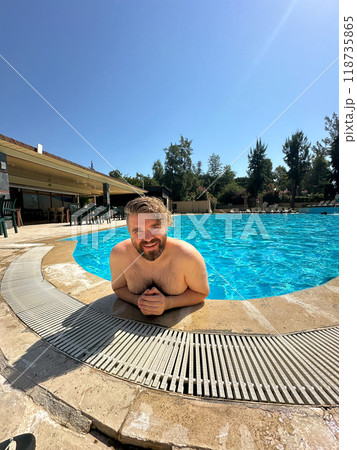 Millennial man enjoying a refreshing day by swimming pool, leaning on the pool's edge and beaming with a broad smile. Vibrant blue water and relaxed setting create a perfect atmosphere for summer fun Millennial man enjoying a refreshing day by swimming pool, leaning on the pool's edge and beaming with a broad smile. Vibrant blue water and relaxed setting create a perfect atmosphere for summer fun 118735865
