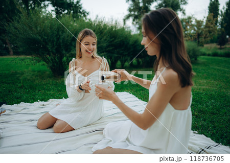 Two friends enjoy a picnic outdoors, sharing drinks in a serene park Two friends enjoy a picnic outdoors, sharing drinks in a serene park 118736705