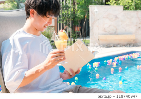 プールサイドでリクライニングチェアに座りトロピカルジュースを飲みながら読書する若いアジア人男性 プールサイドでリクライニングチェアに座りトロピカルジュースを飲みながら読書する若いアジア人男性 118740947