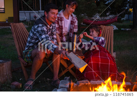 Family is sitting by the outdoor campfire in the courtyard of the house on camping chairs, drink tea from a thermos, warm up and chat by the hearth Family is sitting by the outdoor campfire in the courtyard of the house on camping chairs, drink tea from a thermos, warm up and chat by the hearth 118745865