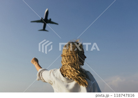 A woman with her arms raised above her head waves at an airplane flying over her 118747816