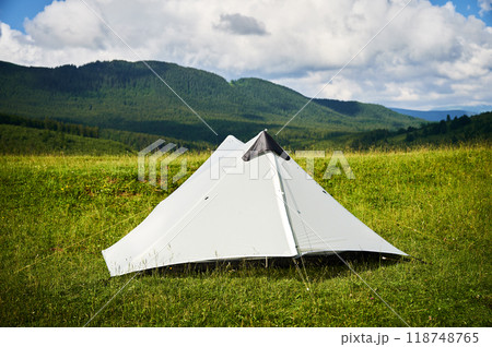 White tourist tent on sunlit, grassy hilltop, surrounded by young pine trees. Panoramic view of lush, rolling mountains under bright blue sky with scattered clouds, creating idyllic camping spot. 118748765