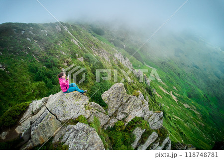 Hiker sits on edge of large rock, overlooking lush, green valley with steep, rocky slopes. Woman taking moment to capture view on phone, surrounded by mist that adds serene, ethereal quality to scene. Hiker sits on edge of large rock, overlooking lush, green valley with steep, rocky slopes. Woman taking moment to capture view on phone, surrounded by mist that adds serene, ethereal quality to scene. 118748781