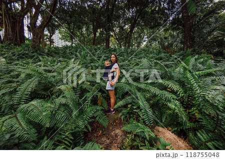 Mother with her daughter in baby carrier in park between fern leaves 118755048