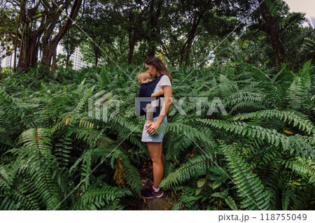 Mother with cute daughter in baby carrier in park between fern leaves 118755049