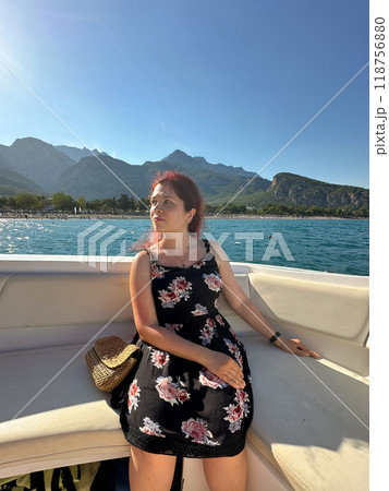 Woman in a black dress sits on a boat under the bright sun, with a scenic view of mountains and the sea in the background. Moment of tranquility and natural beauty concept. Vacation, travel and voyage 118756880