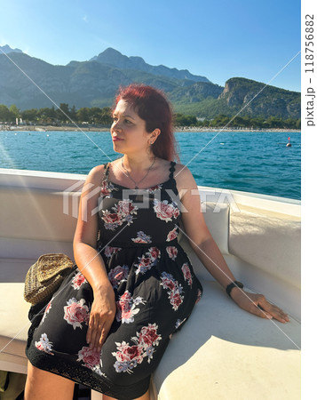 Woman in a black dress sits on a boat under the bright sun, with a scenic view of mountains and the sea in the background. Moment of tranquility and natural beauty concept. Vacation, travel and voyage 118756882