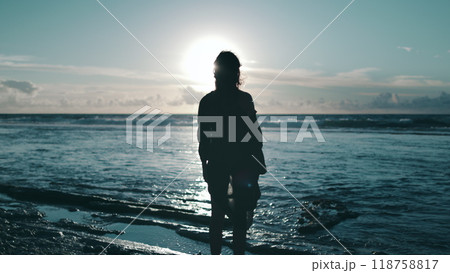 A person stands alone on a sandy beach next to the roaring ocean waves, staring out towards the horizon. The individual appears contemplative, taking in the vast expanse of the sea. 118758817