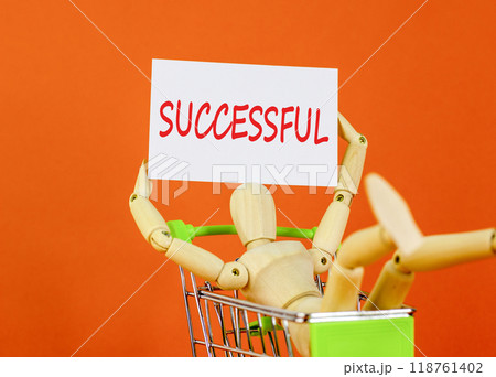 Successful symbol. Concept word Successful on beautiful white paper. Beautiful orange background. Businessman model in shop cart. Business successful concept. Copy space. Successful symbol. Concept word Successful on beautiful white paper. Beautiful orange background. Businessman model in shop cart. Business successful concept. Copy space. 118761402