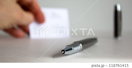 Pen on beautiful white fon on warm light. Fingers with visit cards and cap in the background. 118761415