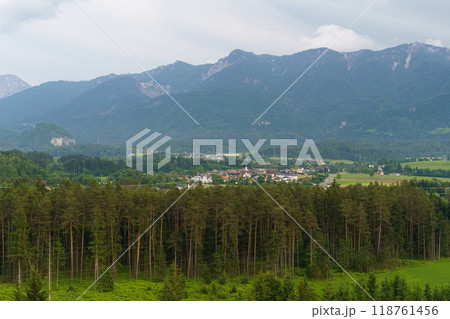 A tranquil village nestled beneath majestic mountains surrounded by lush forests under a cloudy sky in Europe A tranquil village nestled beneath majestic mountains surrounded by lush forests under a cloudy sky in Europe 118761456