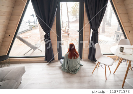 Rear view of a red-haired woman wrapped in a blanket looking out the patio window. Rear view of a red-haired woman wrapped in a blanket looking out the patio window. 118771578