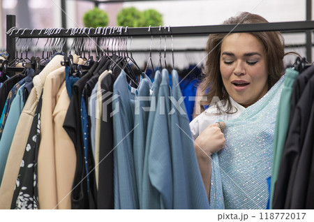 Portrait of a fat woman in a plus size store through hangers with clothes. Portrait of a fat woman in a plus size store through hangers with clothes. 118772017