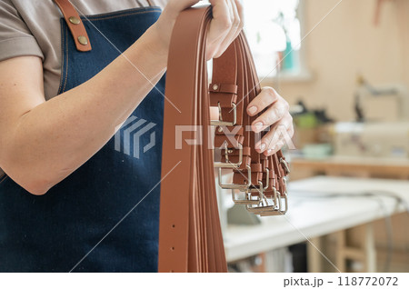 Close-up of a Caucasian woman's hands making genuine leather belts in a workshop. Close-up of a Caucasian woman's hands making genuine leather belts in a workshop. 118772072