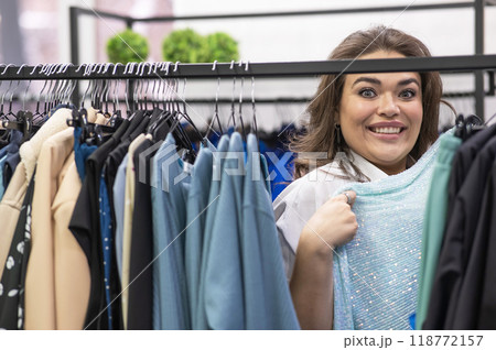 Portrait of a fat woman in a plus size store through hangers with clothes.  118772157