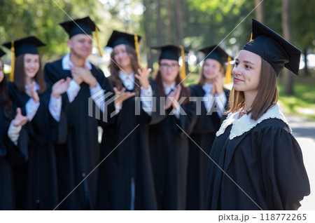 Group of happy students in graduation gowns outdoors. A young girl in the foreground. 118772265
