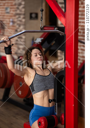 Caucasian forty-year-old woman doing lat pull-downs in the gym. Vertical photo. Caucasian forty-year-old woman doing lat pull-downs in the gym. Vertical photo. 118772329