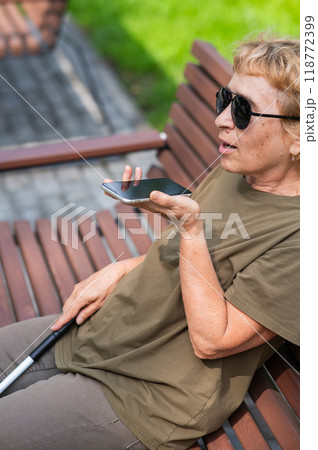 An elderly blind woman sits on a bench in the park and talks on a smartphone. Vertical photo. 118772399