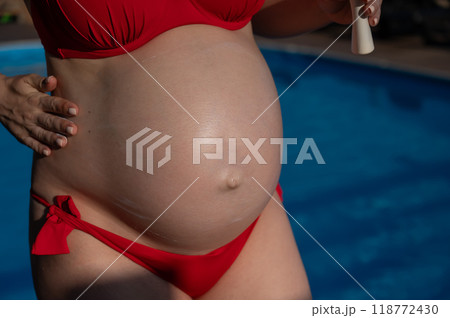 Close-up of the belly of a pregnant woman sunbathing in a red bikini. Expectant mother applies sunscreen.  118772430