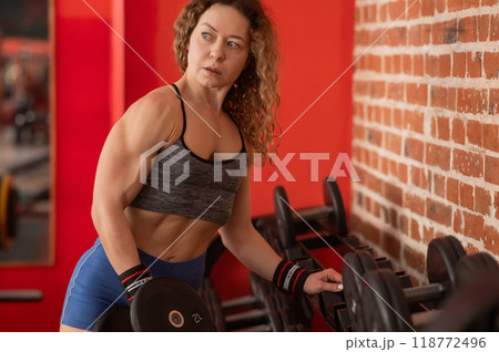Middle aged woman takes dumbbell from rack in gym.  118772496
